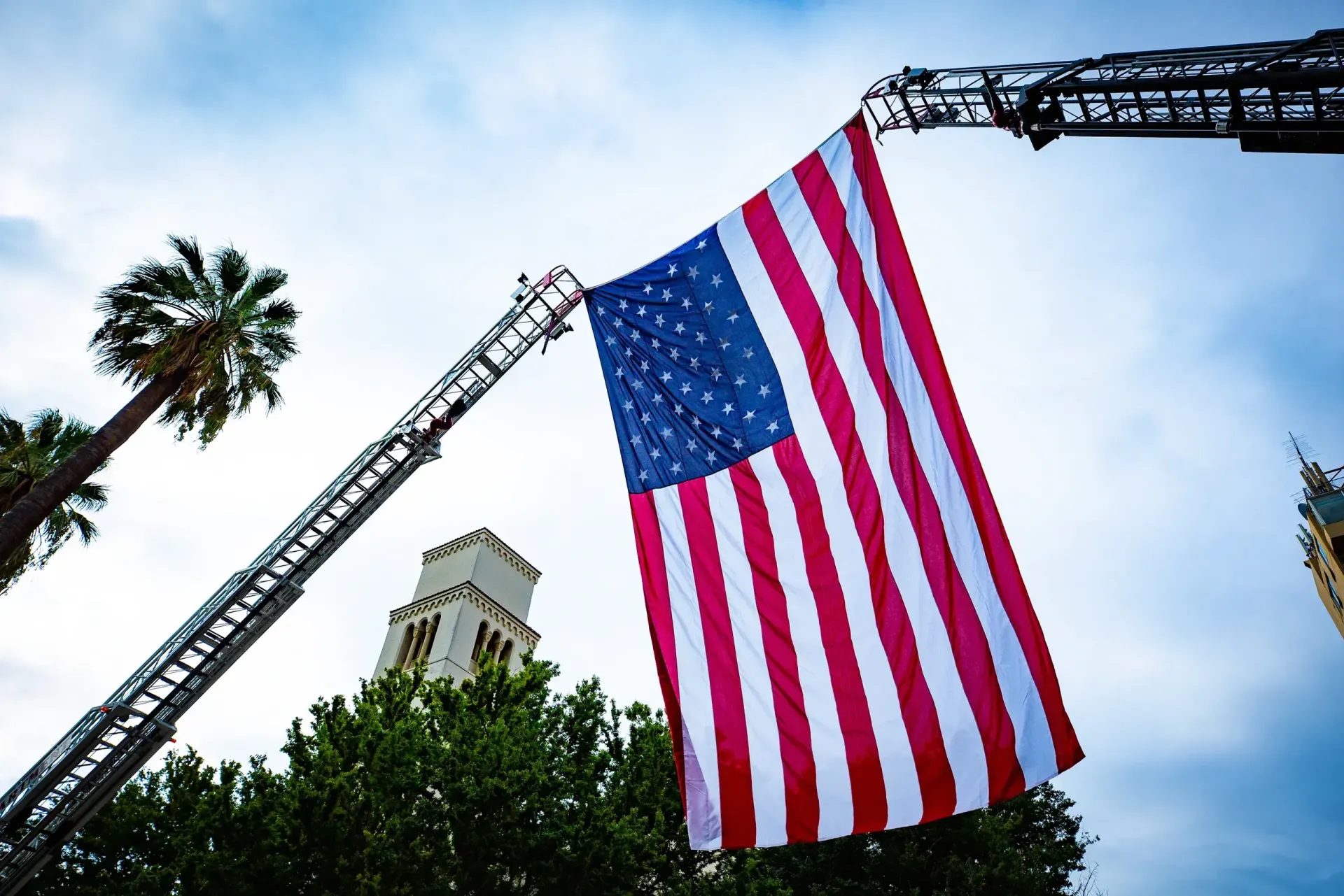A large American flag suspended between two raised fire truck ladders against the sky